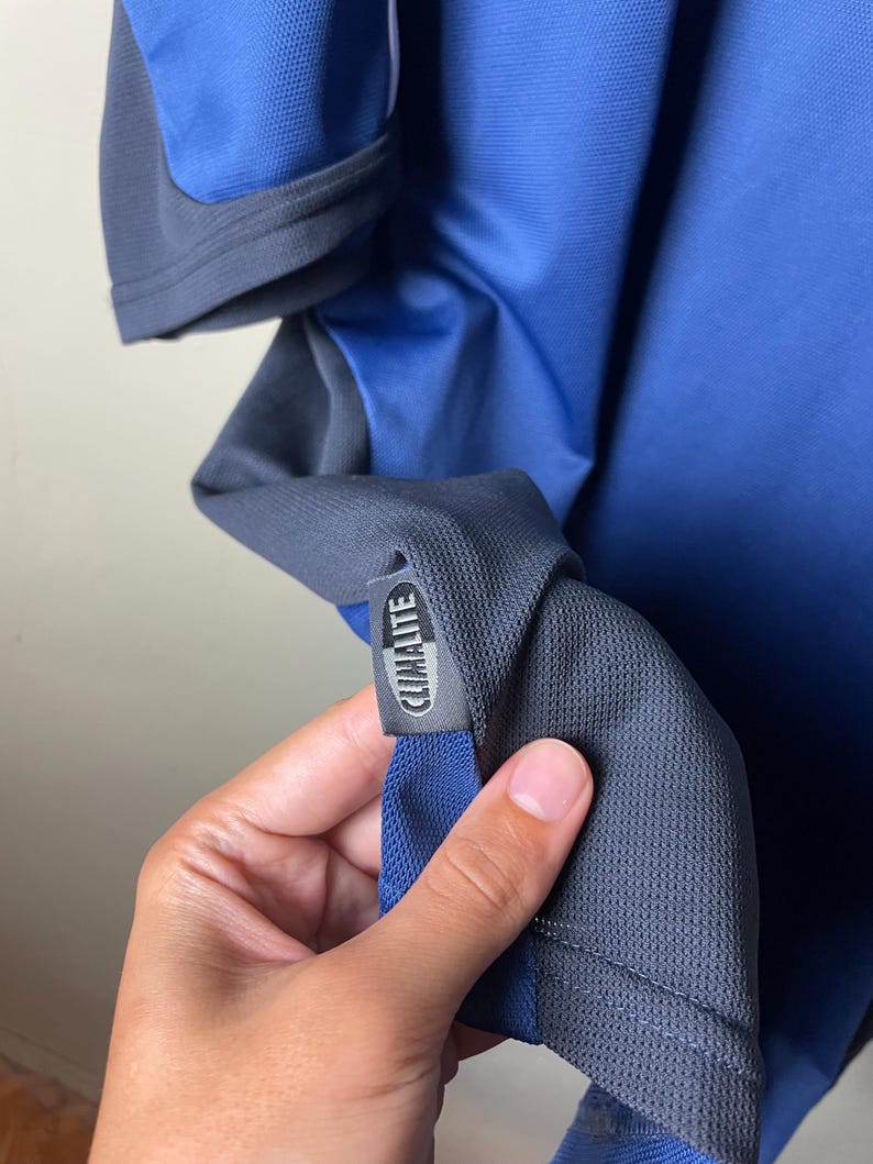 May include: A blue and dark gray athletic shirt featuring a textured mesh fabric. A small rectangular tag is attached to the side seam, displaying the text CLIMALITE in white capital letters against a dark gray background with a blue border.
