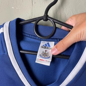 May include: A blue V-neck jersey with white trim along the collar. The interior tag features the Adidas logo above the Newcastle United football club crest. The tag also displays the text Newcastle United and includes a size label of 2XL.