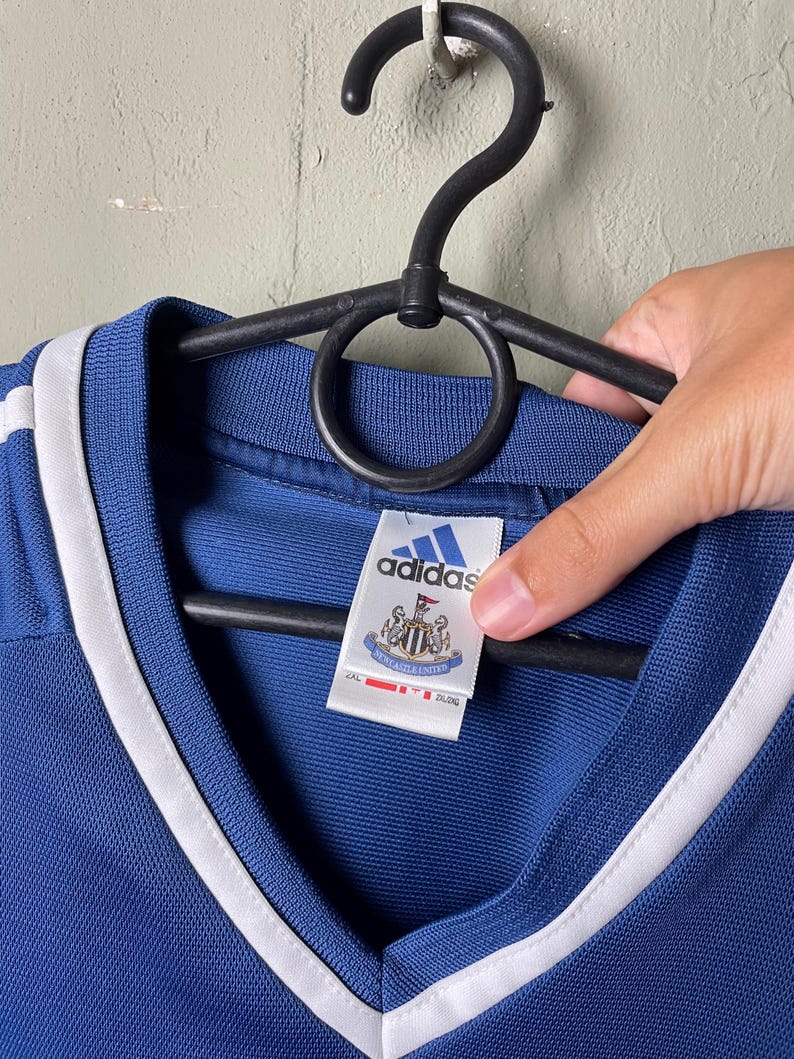 May include: A blue V-neck jersey with white trim along the collar. The interior tag features the Adidas logo above the Newcastle United football club crest. The tag also displays the text Newcastle United and includes a size label of 2XL.