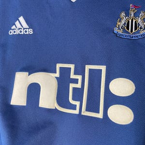 May include: A blue athletic jersey with a white V-neck collar. The front features a white Adidas logo on the upper left, a Newcastle United crest on the upper right, and the white text ntl: printed in a large, bold, sans-serif font across the chest.