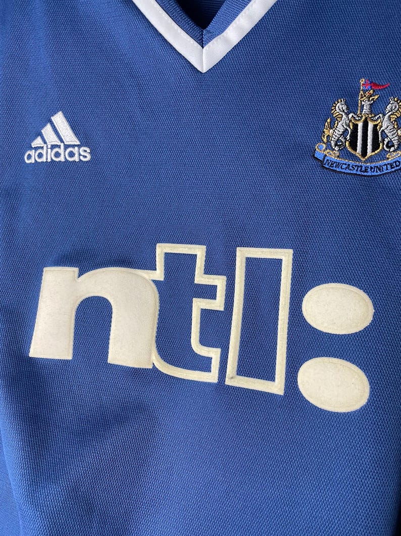 May include: A blue athletic jersey with a white V-neck collar. The front features a white Adidas logo on the upper left, a Newcastle United crest on the upper right, and the white text ntl: printed in a large, bold, sans-serif font across the chest.