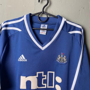 May include: A blue Adidas soccer jersey featuring a V-neck collar with white trim. The Newcastle United crest is on the chest, with the white text ntl: printed below. White stripes run along the shoulders, and an Adidas logo is on the right chest.