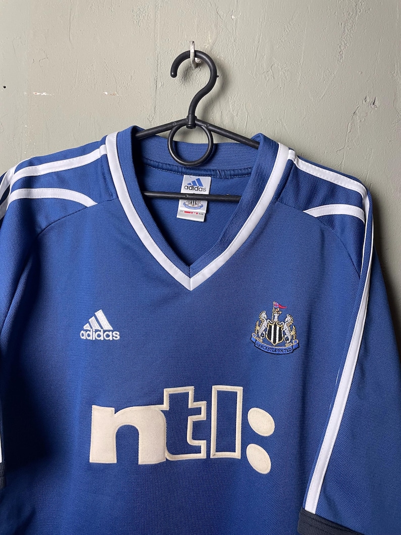 May include: A blue Adidas soccer jersey featuring a V-neck collar with white trim. The Newcastle United crest is on the chest, with the white text ntl: printed below. White stripes run along the shoulders, and an Adidas logo is on the right chest.