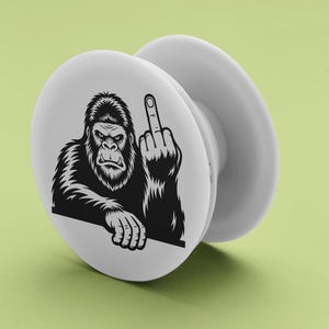 May include: White pop-up phone grip featuring a black and white illustration of a gorilla giving the middle finger. The gorilla is depicted with a stern expression and detailed fur. The grip is round with a collapsible design.
