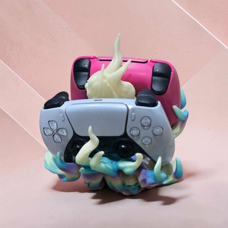 May include: A dual gaming controller stand featuring a sculpted base with swirling flame-like details in pastel blue, purple, and yellow. The stand holds one white and one pink wireless controller, with a central off-white flame accent piece.