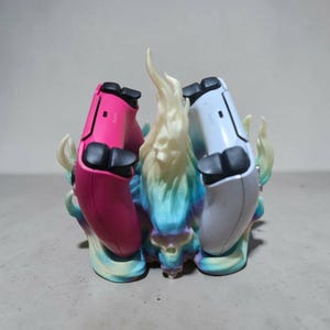 May include: A gaming controller stand featuring a central sculpted skull surrounded by stylized flames in a gradient of blue, purple, and yellow. The stand holds one bright pink controller on the left and one white controller on the right side.