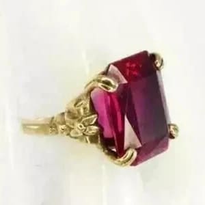 Peut inclure: Une bague dorée ornée d'une grande pierre précieuse rectangulaire rouge foncé. Le sertissage de la bague présente des détails floraux et de feuilles complexes, la pierre étant maintenue par quatre griffes.
