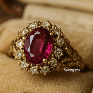 May include: A gold ring featuring a large oval ruby center stone surrounded by small diamonds. The ring has an ornate, floral-inspired design. The ruby is a deep red color, and the diamonds sparkle against the gold setting. The ring is displayed in a velvet jewelry box.