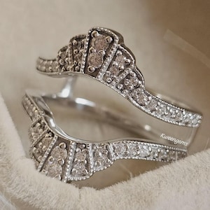 Könnte beinhalten: Zwei silberfarbene Ringe mit funkelndem Design. Jeder Ring hat eine Reihe kleiner, klarer Steine und eine dekorative Oberseite mit größeren Steinen. Die Ringe sind vor einem weichen, weißen Hintergrund platziert.