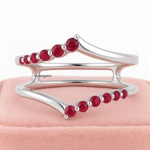 May include: A silver ring with a curved design, featuring a row of small, round, red gemstones. The ring has a double-band design, with the gemstones set along the top and bottom curves. The ring is displayed on a pink surface.