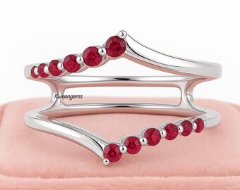 May include: A silver ring with a curved design, featuring a row of small, round, red gemstones. The ring has a double-band design, with the gemstones set along the top and bottom curves. The ring is displayed on a pink surface.