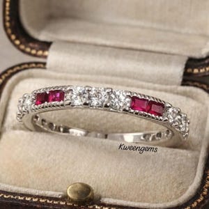 May include: A silver ring with alternating round white gemstones and square red gemstones. The ring is set in a vintage-style box. The text "Kweengems" is visible on the ring.