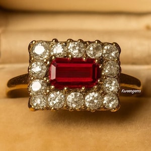 May include: A gold ring with a central, rectangular, deep red gemstone, framed by a square of smaller, clear gemstones. The ring's band is plain, polished gold.