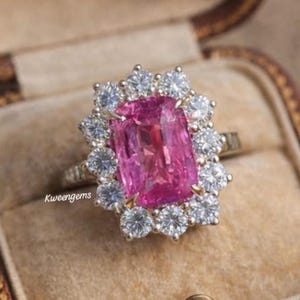 Peut inclure: Une bague ornée d'une pierre rose rectangulaire, entourée d'un halo de petites pierres claires. La bague est sertie dans un anneau en or. Le texte "Kweengems" est visible sur la bague.