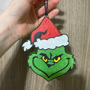 May include: A green Grinch-themed ornament with a red Santa hat and black string for hanging. The Grinch's face is detailed with yellow eyes and a black outline, perfect for holiday decor.