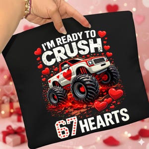 Pode incluir: Almofada preta com um gráfico de um monster truck vermelho e branco com um coração. O texto diz "I'M READY TO CRUSH" e "67 HEARTS" com detalhes de coração vermelho. Um tema divertido do Dia dos Namorados.