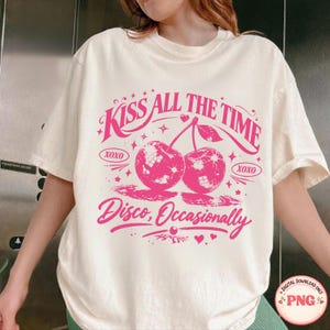 May include: A cream-coloured t-shirt with a pink graphic. The graphic features the text "Kiss All The Time" above two disco ball cherries and the phrase "Disco, Occasionally" below. The design includes hearts and stars.