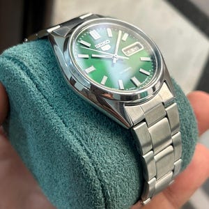 May include: A stainless steel Seiko 5 automatic watch with a green dial. The watch features a silver band and a day-date display. The watch is displayed on a teal-colored cushion.