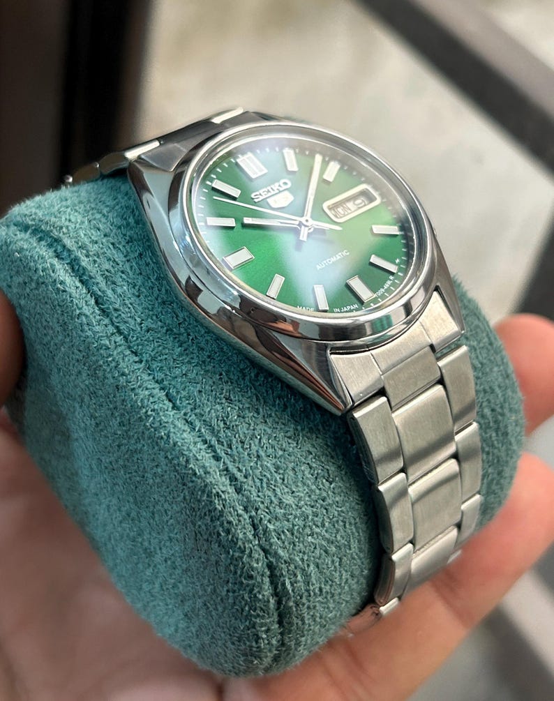 May include: A stainless steel Seiko 5 automatic watch with a green dial. The watch features a silver band and a day-date display. The watch is displayed on a teal-colored cushion.
