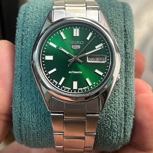 May include: A stainless steel Seiko 5 automatic watch with a vibrant green dial. The watch features a silver bezel, a metal band, and a day-date display. The dial includes the Seiko logo and the word "AUTOMATIC".
