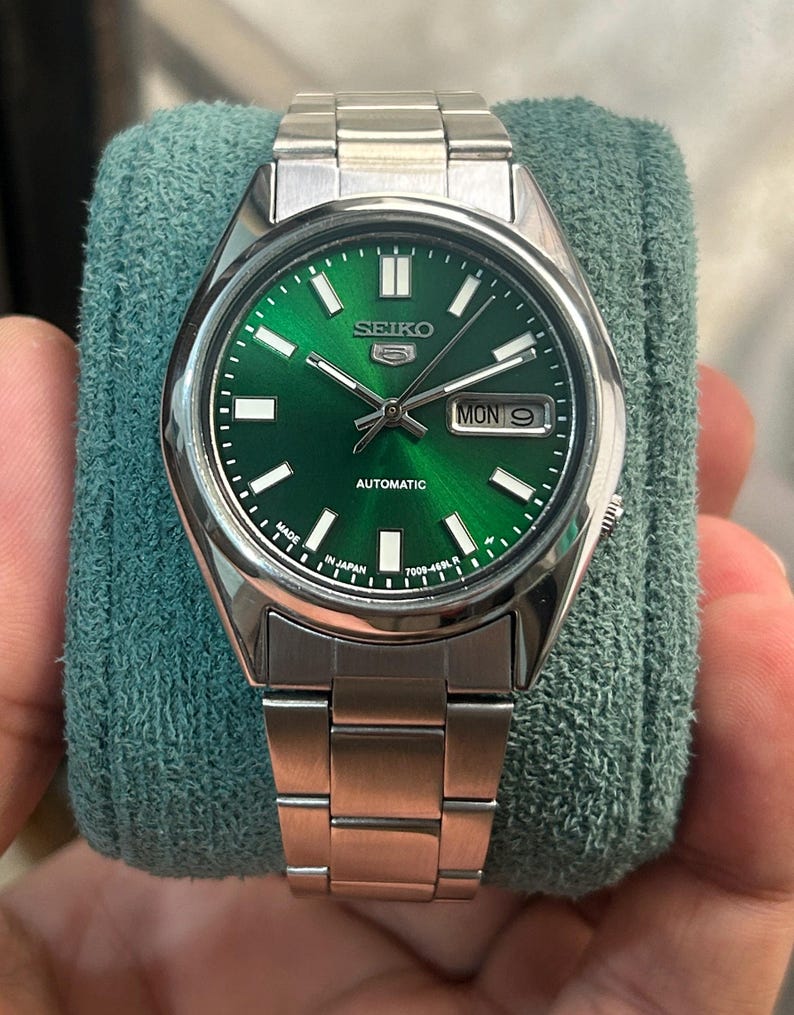 May include: A stainless steel Seiko 5 automatic watch with a vibrant green dial. The watch features a silver bezel, a metal band, and a day-date display. The dial includes the Seiko logo and the word "AUTOMATIC".