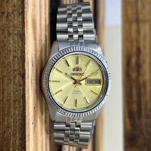 May include: A silver-toned Orient watch with a gold-colored face and a stainless steel band. The watch face displays the day and date, with the day reading "TUE 30". The watch has a fluted bezel and a crystal face.