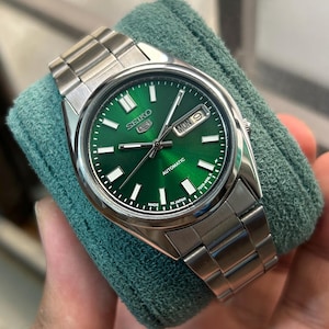 May include: A stainless steel Seiko 5 automatic watch with a green dial. The watch features a day and date display, luminous hands and markers, and a stainless steel bracelet. The watch is displayed on a green cloth.