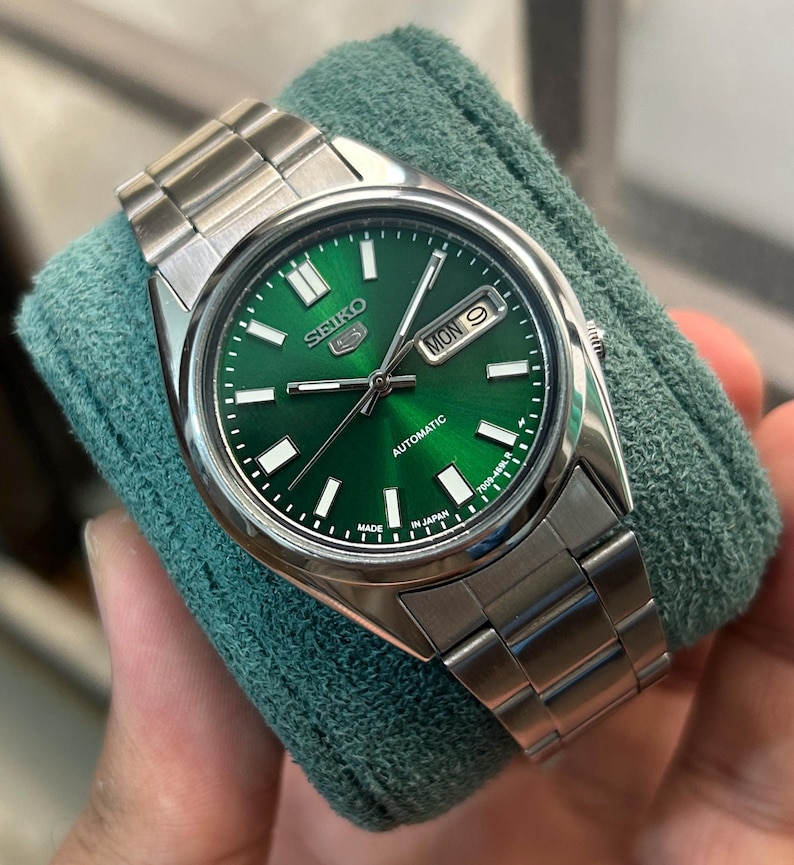 May include: A stainless steel Seiko 5 automatic watch with a green dial. The watch features a day and date display, luminous hands and markers, and a stainless steel bracelet. The watch is displayed on a green cloth.