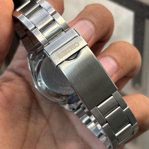 May include: Close-up of a stainless steel Seiko watch. The watch features a silver metal band with a clasp that has the Seiko logo. The watch face is visible, with a silver bezel and a dark-colored dial.
