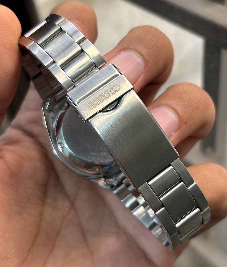 May include: Close-up of a stainless steel Seiko watch. The watch features a silver metal band with a clasp that has the Seiko logo. The watch face is visible, with a silver bezel and a dark-colored dial.