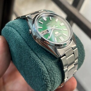 May include: A stainless steel Seiko automatic watch with a green dial. The watch features a silver band and a date display. The watch is displayed on a teal green velvet surface.