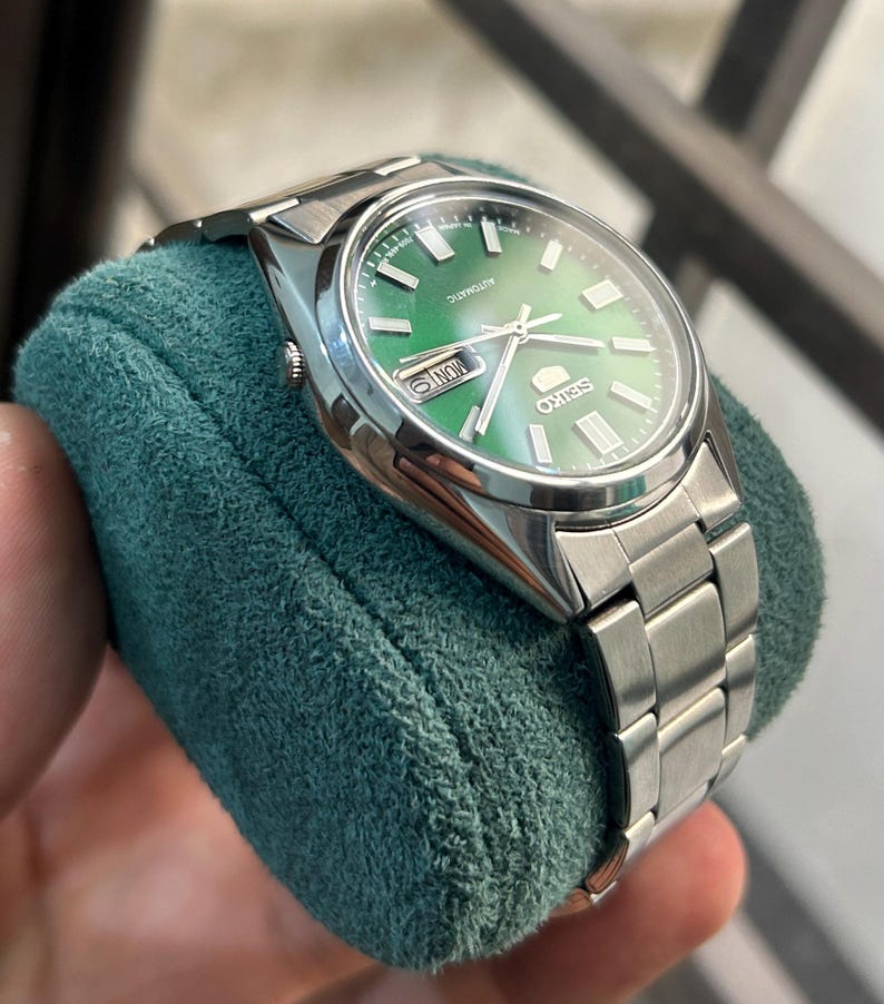May include: A stainless steel Seiko automatic watch with a green dial. The watch features a silver band and a date display. The watch is displayed on a teal green velvet surface.