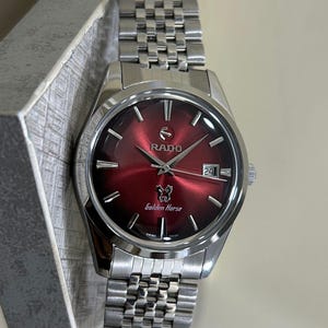 May include: A stainless steel Rado Golden Horse watch with a red dial. The watch features a date window at the 3 o'clock position and a silver-tone bracelet. The watch is displayed in a gray box.