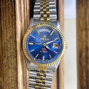 May include: A silver and gold wristwatch with a blue face. The watch face displays "Saturday" and the brand name "Ricoh". The watch has a gold bezel and a silver and gold band. The watch is likely made in Japan.