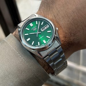 May include: A stainless steel Seiko automatic watch with a vibrant green dial. The watch features a date display and a silver band. The watch is displayed on a wrist, showcasing the watch's design and details.