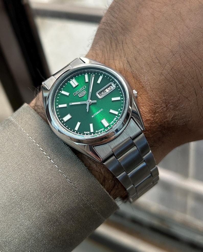 May include: A stainless steel Seiko automatic watch with a vibrant green dial. The watch features a date display and a silver band. The watch is displayed on a wrist, showcasing the watch's design and details.