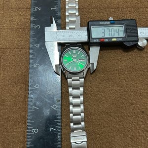 May include: A silver Seiko watch with a green dial, measured at 37.04 mm. The watch has a stainless steel band and is being measured with a digital caliper and a ruler. The watch face displays the day and date.