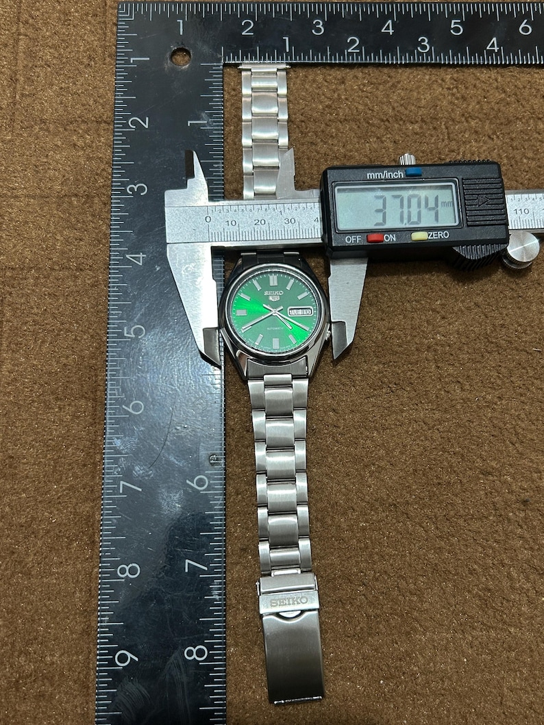 May include: A silver Seiko watch with a green dial, measured at 37.04 mm. The watch has a stainless steel band and is being measured with a digital caliper and a ruler. The watch face displays the day and date.