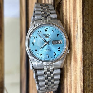 Puede incluir: Un reloj de pulsera Seiko plateado con esfera azul claro y números arábigos. El reloj tiene una pantalla de fecha y una correa de eslabones plateados. El reloj está etiquetado como "Automatic 17 Jewels".