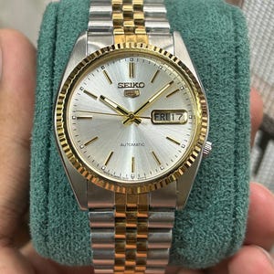 May include: A Seiko 5 automatic wristwatch featuring a silver sunray dial with gold-tone indices and hands. The watch has a fluted gold-tone bezel and a two-tone stainless steel jubilee-style bracelet with gold-tone center links and a day-date window.