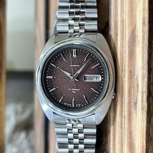 Może przedstawiać: Zegarek automatyczny Seiko w stylu vintage z brązową tarczą i srebrnymi wskazówkami i znacznikami. Zegarek posiada wyświetlacz dnia i daty oraz bransoletę ze stali nierdzewnej. Zegarek jest prezentowany na drewnianym tle.