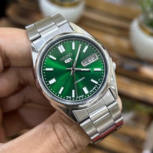 May include: A stainless steel wristwatch with a vibrant green dial. The watch features white hour markers, a day-date display, and a silver-tone metal band. The watch face displays the brand name "Seiko" and the word "Automatic".