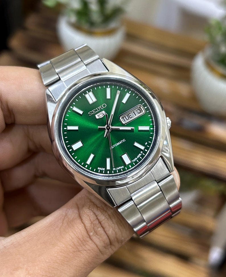 May include: A stainless steel wristwatch with a vibrant green dial. The watch features white hour markers, a day-date display, and a silver-tone metal band. The watch face displays the brand name "Seiko" and the word "Automatic".