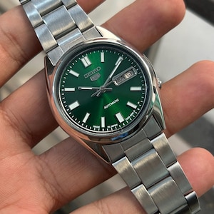May include: A stainless steel Seiko automatic wristwatch with a vibrant green dial. The watch features a day and date display, luminous hands and markers, and a silver-tone bracelet. The watch face displays the text "SEIKO" and "AUTOMATIC".