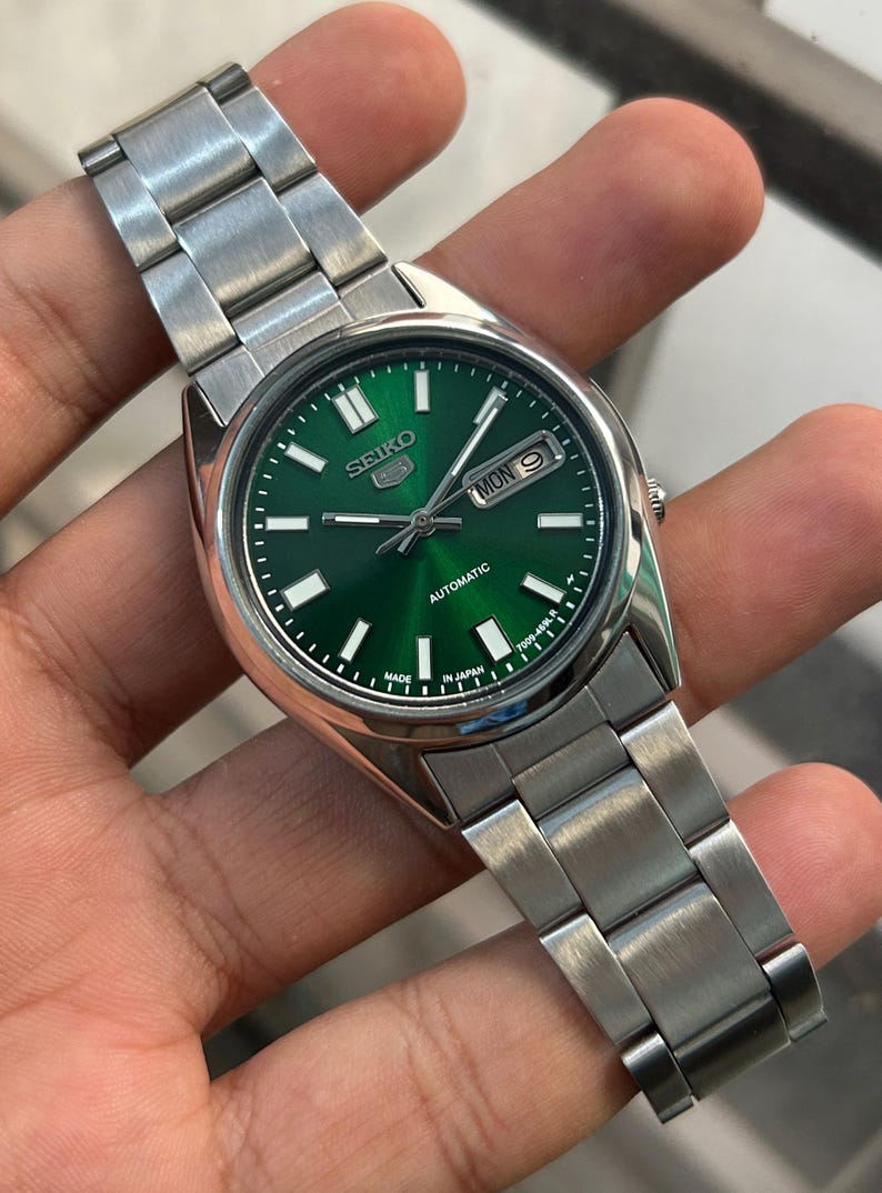 May include: A stainless steel Seiko automatic wristwatch with a vibrant green dial. The watch features a day and date display, luminous hands and markers, and a silver-tone bracelet. The watch face displays the text "SEIKO" and "AUTOMATIC".