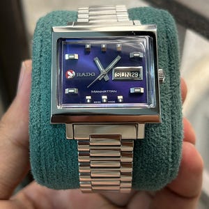 Puede incluir: Un reloj Rado Manhattan plateado con esfera azul cuadrada. El reloj tiene una correa plateada y muestra el día como domingo 29. El reloj es de fabricación suiza.