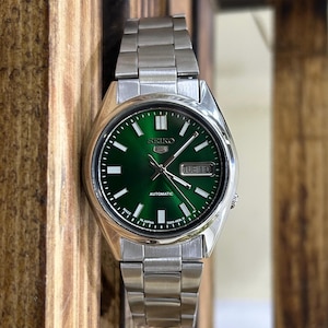 May include: A stainless steel Seiko automatic watch with a vibrant green dial. The watch features a day and date display, luminous hands and markers, and a silver-tone bracelet. The watch is made in Japan.