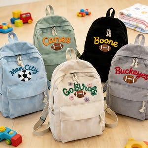 Może przedstawiać: Pięć sztruksowych plecaków w różnych kolorach: jasnoniebieskim, zielonym, czarnym, szarym i kremowym. Każdy plecak ma haftowany tekst i grafikę sportową, w tym piłkę nożną lub futbol amerykański. Plecaki mają przednią kieszeń i regulowane paski.