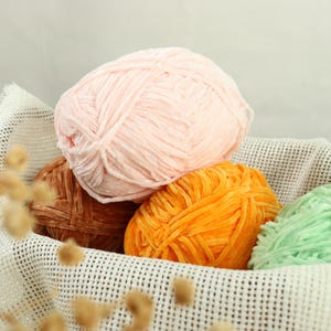 Puede incluir: Una cesta llena de ovillos de lana coloridos. La lana es en tonos rosa, naranja, marrón y verde. La lana tiene una textura suave y aterciopelada. La cesta es blanca y tiene un patrón de malla.