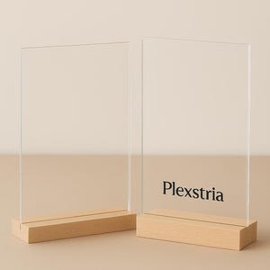 May include: Two clear acrylic sign holders with wooden bases. The sign holders are rectangular and stand upright. One sign holder has the word "Plexstria" printed in black. The background is a neutral beige.